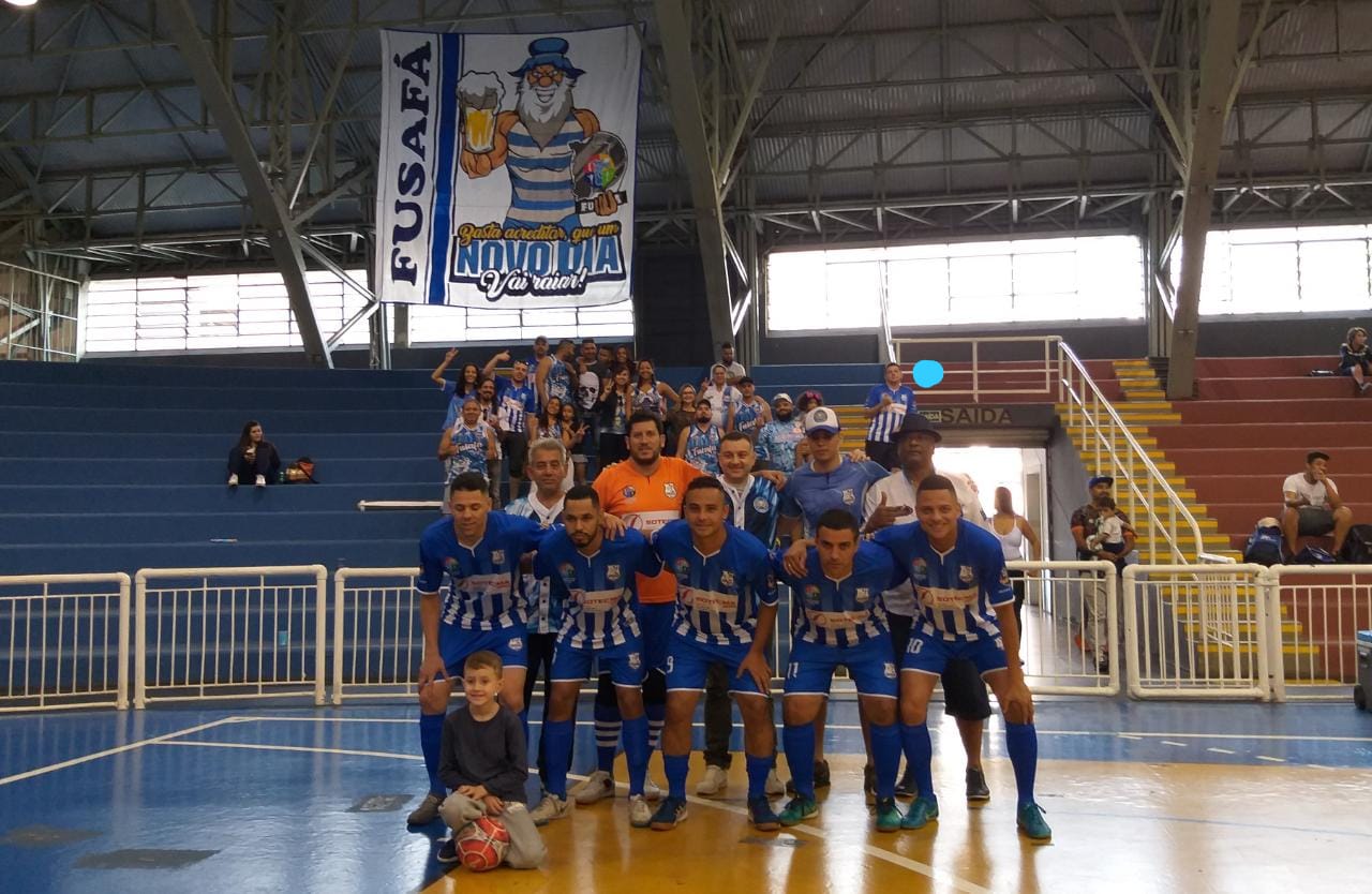 Fotografia colorida mostra quadra de futsal com time Fusafá em pose para foto. Todos vestem uniforme azul 